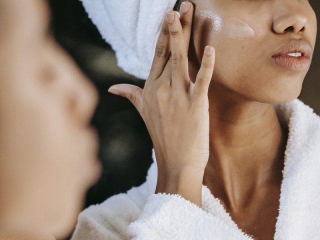 Crop ethnic woman applying cream on face against mirror